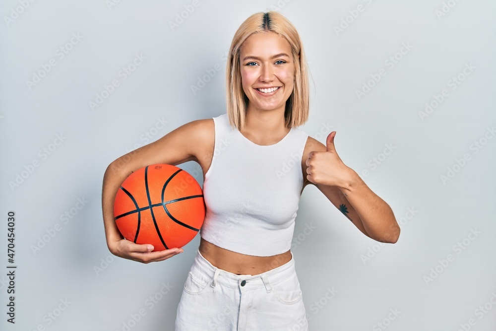 Beautiful blonde woman holding basketball ball smiling happy and positive, thumb up doing excellent and approval sign
