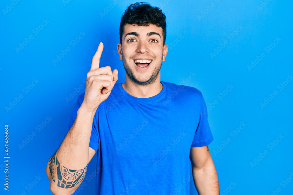 Fototapeta premium Young hispanic man wearing casual blue t shirt pointing finger up with successful idea. exited and happy. number one.