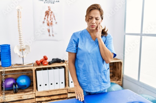 Middle age hispanic physiotherapist woman working at pain recovery clinic touching mouth with hand with painful expression because of toothache or dental illness on teeth. dentist