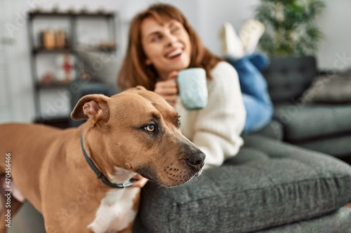 Young caucasian woman drinking coffee lying on sofa with dog at home