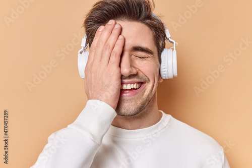 Positive guy covers face with hand keeps eyes closed smiles gladfully listens audio track via headphones wears white jumper isolated over beige background. People emotions and hobby concept.