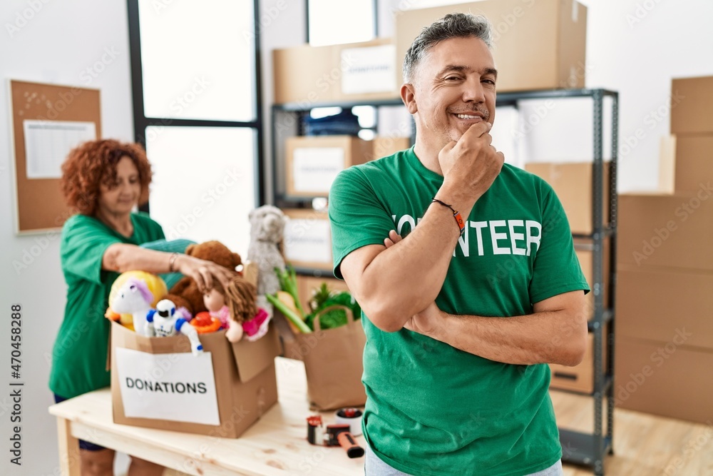 Middle age man wearing volunteer t shirt at donations stand looking confident at the camera smiling with crossed arms and hand raised on chin. thinking positive. Middle age man wearing volunteer t shirt at donations stand looking confident at the camera smiling with crossed arms and hand raised on chin. thinking positive.