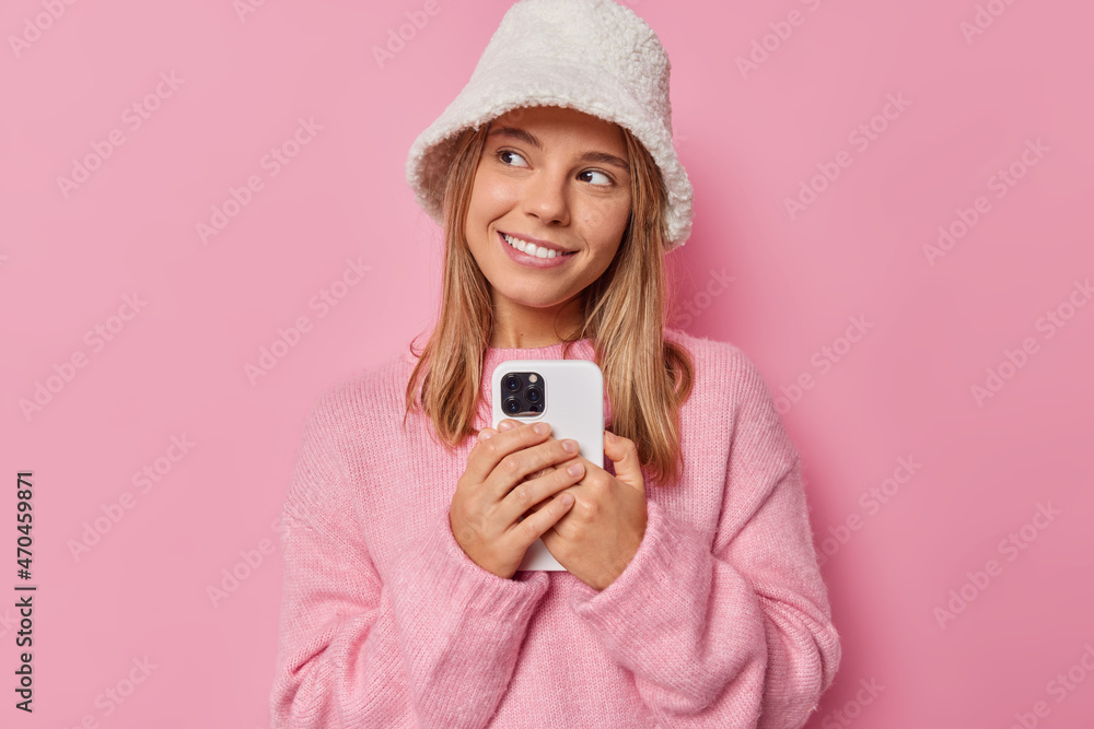 Horizontal shot of beautiful young woman with dreamy expression looks away smiles gladfully holds mobile phone thinks what answer to give on received message wears casual jumper and stylish hat Horizontal shot of beautiful young woman with dreamy expression looks away smiles gladfully holds mobile phone thinks what answer to give on received message wears casual jumper and stylish hat