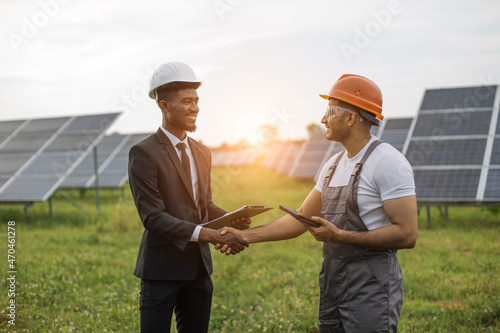 African american man in suit and white helmet shaking hands with indian man in uniform and yellow hard hat. Inspector and engineer standing together among solar plant. © sofiko14