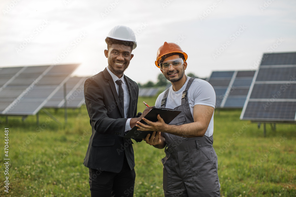 African american man in black suit and white helmet signing papers on clipboard that holding indian man in uniform. Owner and worker standing together among photovoltaic cells. African american man in black suit and white helmet signing papers on clipboard that holding indian man in uniform. Owner and worker standing together among photovoltaic cells.