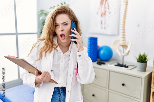Young caucasian woman working at rehabilitation clinic speaking on the phone in shock face  looking skeptical and sarcastic  surprised with open mouth