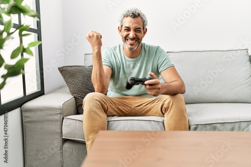 Middle age hispanic man playing video game sitting on the sofa screaming proud, celebrating victory and success very excited with raised arms © Krakenimages.com