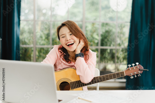 Asian girl is learning and practicing how to play the guitar online at home with her laptop computer. A cheerful female artist plays a song while watching an online training