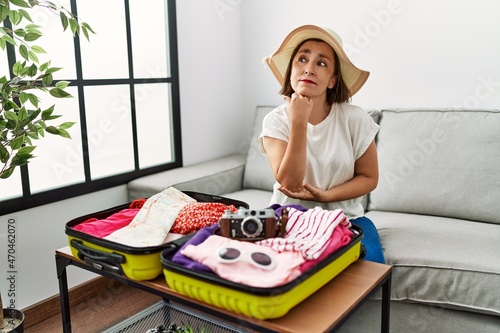 Obraz na plátne Beautiful middle age hispanic woman packing summer clothes in suitcase with hand on chin thinking about question, pensive expression