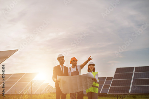 Multi ethnic people in hardhats standing outdoors among solar cells and having working meeting. Competent workers discussing business plant of producing alternative energy.