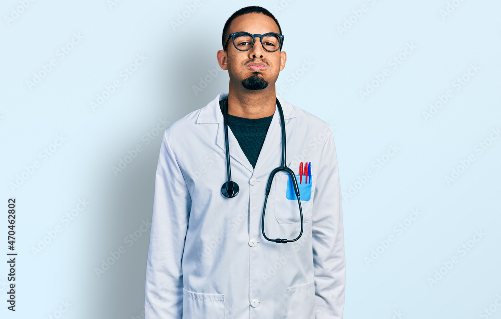 Fototapeta premium Young african american man wearing doctor uniform and stethoscope puffing cheeks with funny face. mouth inflated with air, crazy expression.