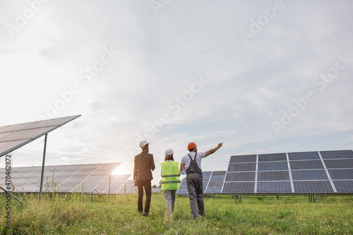 Back view of two inspectors in white helmets walking on solar station with technician in uniform. Multiracial engineers examining work of photovoltaic cells outdoors.