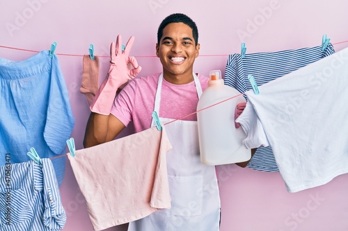 Young handsome hispanic man doing laundry holding detergent bottle doing ok sign with fingers, smiling friendly gesturing excellent symbol