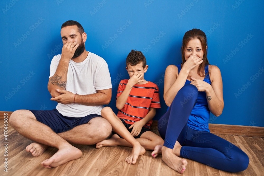 Family of three sitting on the floor at home smelling something stinky and disgusting, intolerable smell, holding breath with fingers on nose. bad smell Family of three sitting on the floor at home smelling something stinky and disgusting, intolerable smell, holding breath with fingers on nose. bad smell