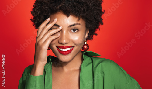 New Year and women beauty. Smiling black woman with red lips and makeup, touching her clean perfect skin, wearing Chistmas toy earrings and green blouse, winter holidays concept