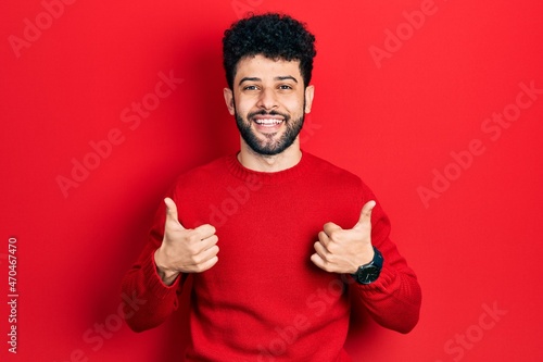 Young arab man with beard wearing casual red sweater success sign doing positive gesture with hand, thumbs up smiling and happy. cheerful expression and winner gesture.