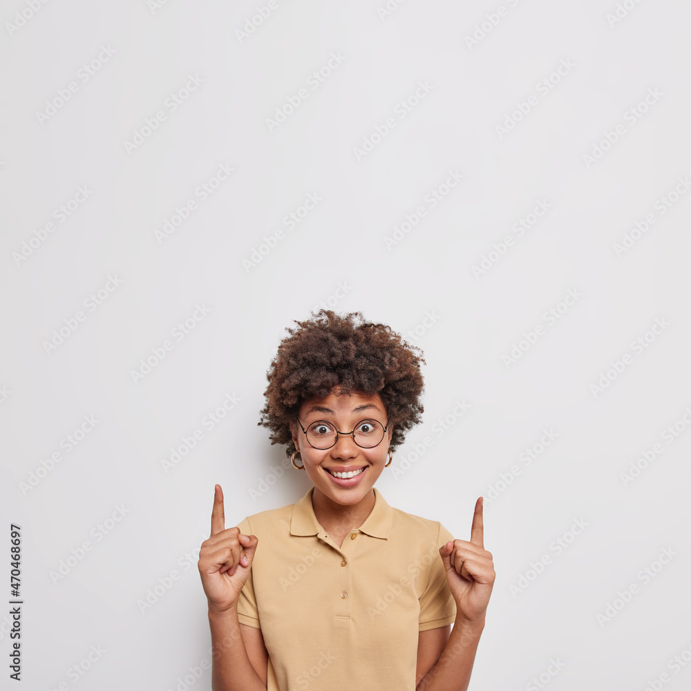Look overhead. Positive woman with cheerful expression points index fingers at blank copy space presents product attracts your attention to promotion wears casual clothes isolated on white background Look overhead. Positive woman with cheerful expression points index fingers at blank copy space presents product attracts your attention to promotion wears casual clothes isolated on white background