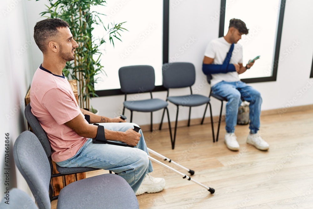 Two hispanic men using smartphone sitting on chair at hospital waiting room