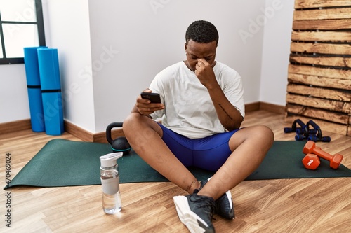 Young african man sitting on training mat at the gym using smartphone tired rubbing nose and eyes feeling fatigue and headache. stress and frustration concept.