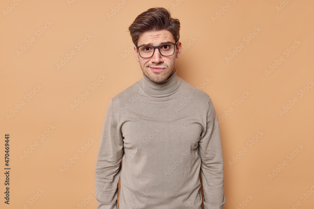 Puzzled displeased man with hairstyle looks unhappily at camera reacts on bad news wears transparent eyeglasses and casual poloneck isolated over beige background. Negative face expressions. Puzzled displeased man with hairstyle looks unhappily at camera reacts on bad news wears transparent eyeglasses and casual poloneck isolated over beige background. Negative face expressions.