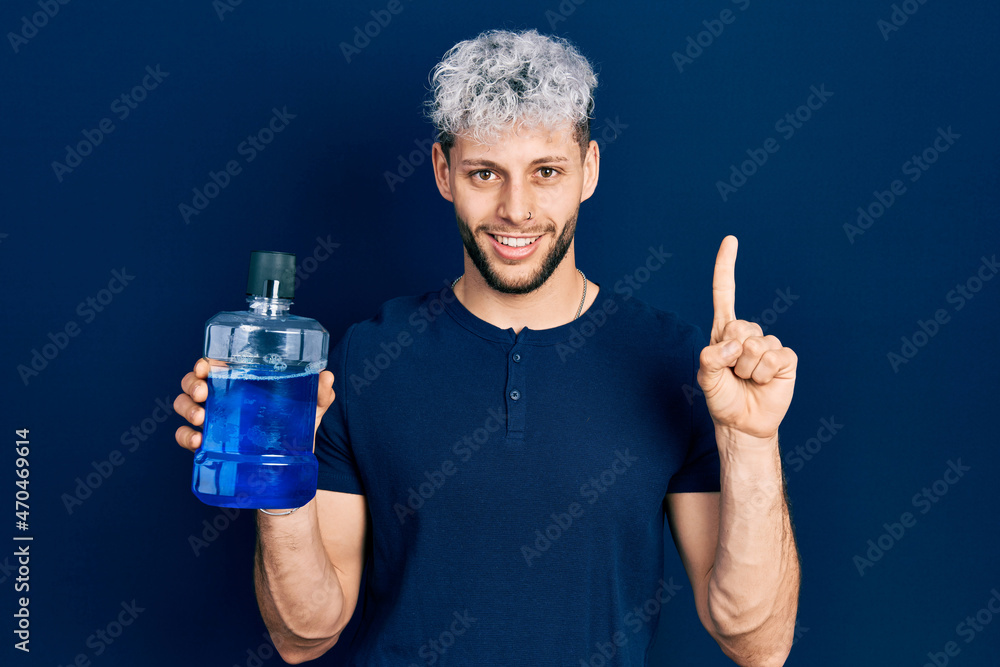 Young hispanic man with modern dyed hair holding mouthwash for fresh breath smiling with an idea or question pointing finger with happy face, number one