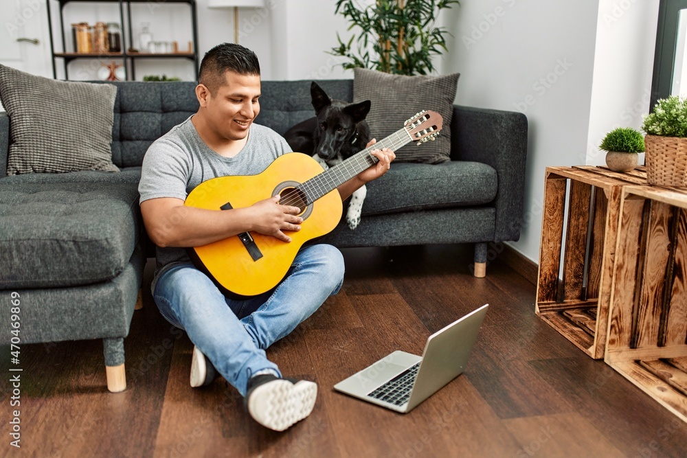 Young hispanic man having onlinr classical guitar lesson sitting on the floor with dog at home.