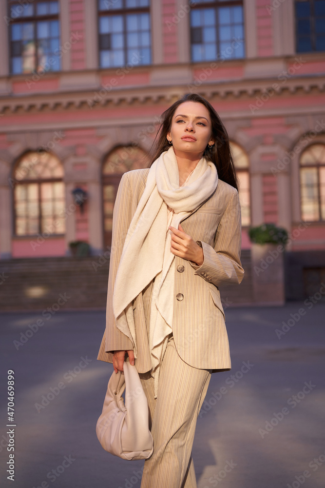 Young beautiful elegant woman outdoor portrait. Fashionable girl in beige blazer, trousers, white shoes and scarf. Lady with long brunette straight hair walking at city street