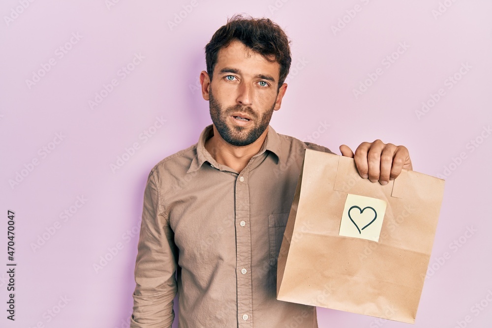 Handsome man with beard holding delivery paper bag with heart reminder looking sleepy and tired, exhausted for fatigue and hangover, lazy eyes in the morning. Handsome man with beard holding delivery paper bag with heart reminder looking sleepy and tired, exhausted for fatigue and hangover, lazy eyes in the morning.