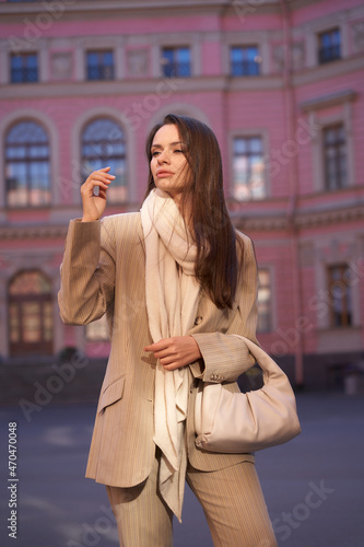 Young beautiful elegant woman outdoor portrait. Fashionable girl in beige blazer, trousers, white shoes and scarf. Lady with long brunette straight hair walking at city street