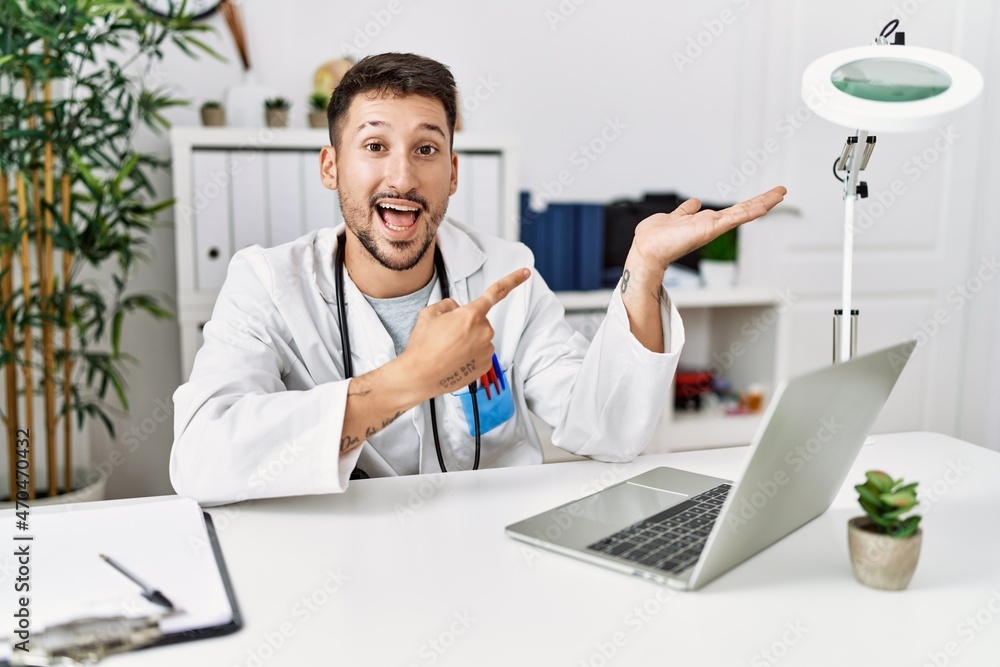 Young doctor working at the clinic using computer laptop amazed and smiling to the camera while presenting with hand and pointing with finger.