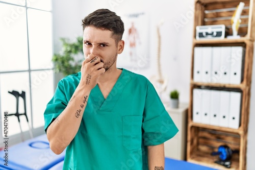 Young physiotherapist man working at pain recovery clinic smelling something stinky and disgusting, intolerable smell, holding breath with fingers on nose. bad smell