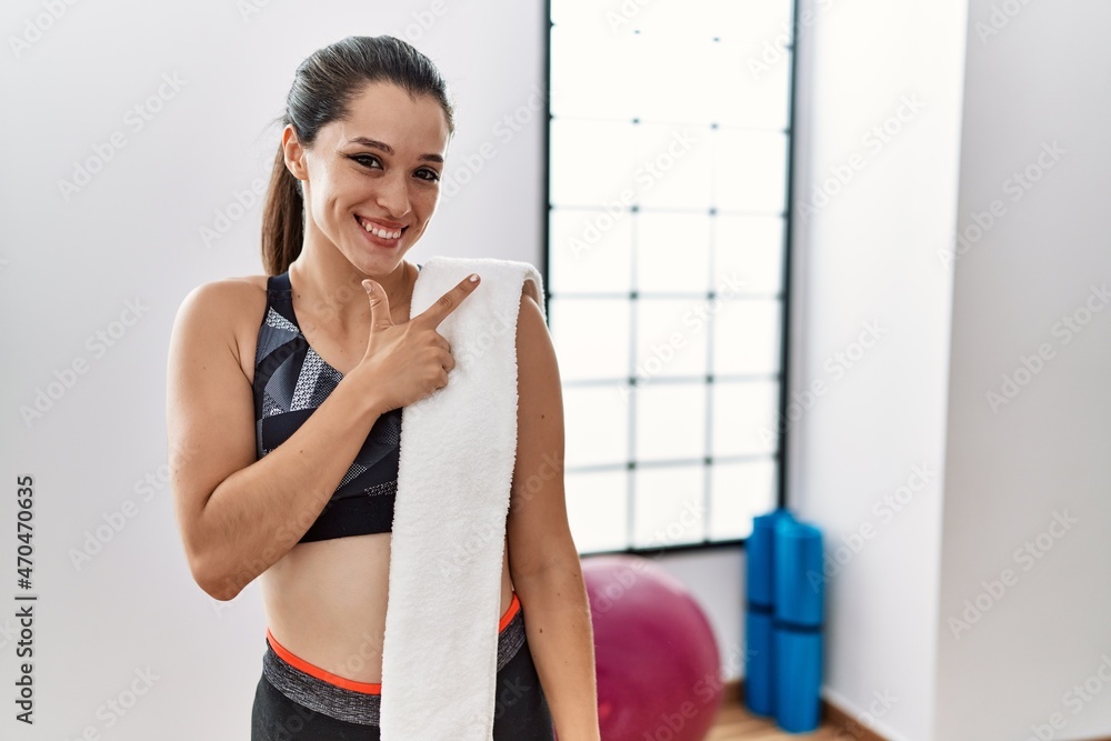 Young brunette woman wearing sportswear and towel at the gym cheerful with a smile on face pointing with hand and finger up to the side with happy and natural expression Young brunette woman wearing sportswear and towel at the gym cheerful with a smile on face pointing with hand and finger up to the side with happy and natural expression