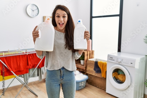 Young brunette woman holding detergent bottle at laundry room celebrating crazy and amazed for success with open eyes screaming excited. photo