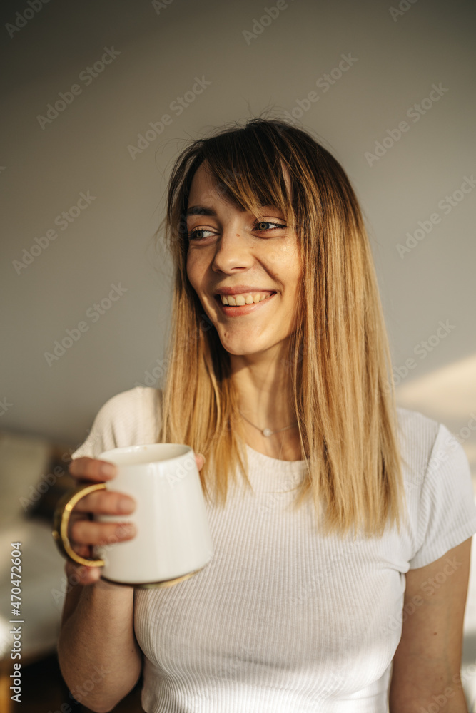 Attractive girl stands in the room and holds a cup of coffee. The girl enjoys the sun. Portrait of a girl with coffee Attractive girl stands in the room and holds a cup of coffee. The girl enjoys the sun. Portrait of a girl with coffee