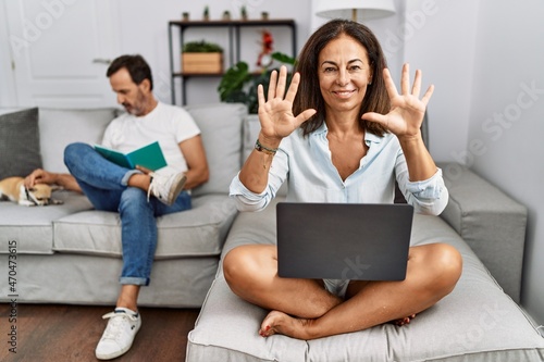 Hispanic middle age couple at home, woman using laptop showing and pointing up with fingers number ten while smiling confident and happy.