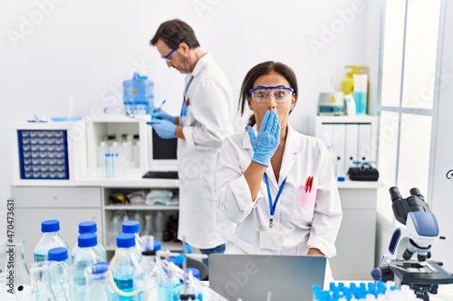 Middle age woman working at scientist laboratory covering mouth with hand, shocked and afraid for mistake. surprised expression