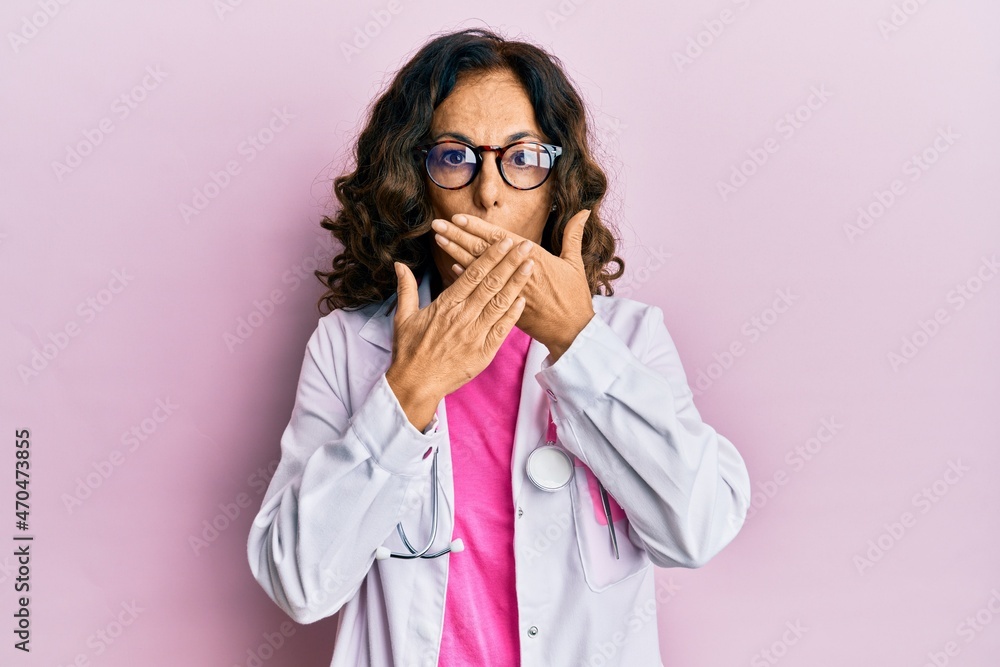 Middle age hispanic woman wearing doctor uniform and glasses shocked covering mouth with hands for mistake. secret concept. Middle age hispanic woman wearing doctor uniform and glasses shocked covering mouth with hands for mistake. secret concept.