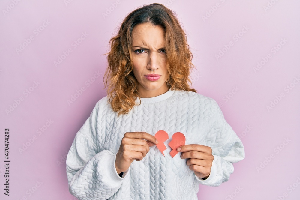 Young caucasian woman holding broken heart paper shape skeptic and nervous, frowning upset because of problem. negative person. Young caucasian woman holding broken heart paper shape skeptic and nervous, frowning upset because of problem. negative person.