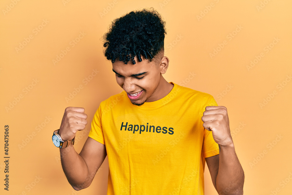Young african american man wearing t shirt with happiness word message very happy and excited doing winner gesture with arms raised, smiling and screaming for success. celebration concept. Young african american man wearing t shirt with happiness word message very happy and excited doing winner gesture with arms raised, smiling and screaming for success. celebration concept.