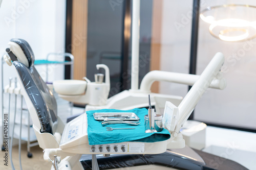 Side view selective focus of dental instruments set placed on a medical disposable blue napkin on an operator s table near an empty dental chair with a blurred bright dental clinic room in background.