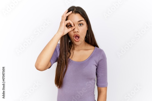 Young brunette woman standing by isolated background doing ok gesture shocked with surprised face, eye looking through fingers. unbelieving expression.