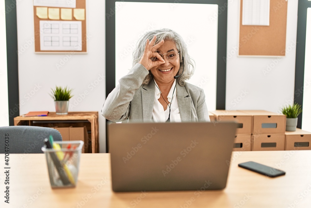 Middle age businesswoman sitting on desk working using laptop at office doing ok gesture with hand smiling, eye looking through fingers with happy face. Middle age businesswoman sitting on desk working using laptop at office doing ok gesture with hand smiling, eye looking through fingers with happy face.