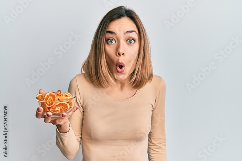 Young caucasian blonde woman holding bowl of dry orange scared and amazed with open mouth for surprise  disbelief face