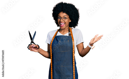 Young african american woman dressmaker designer wearing atelier apron holding scissors celebrating achievement with happy smile and winner expression with raised hand