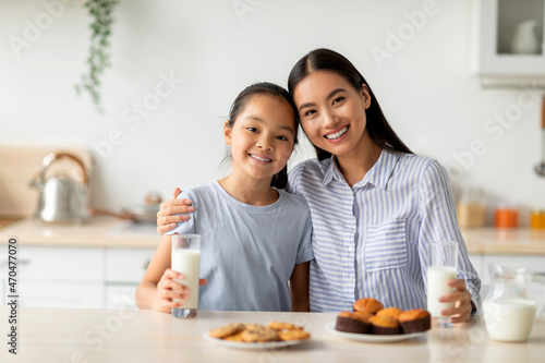Young asian mother and daughter enjoying fresh homemade cookies, drinking milk while sitting at table in kitchen