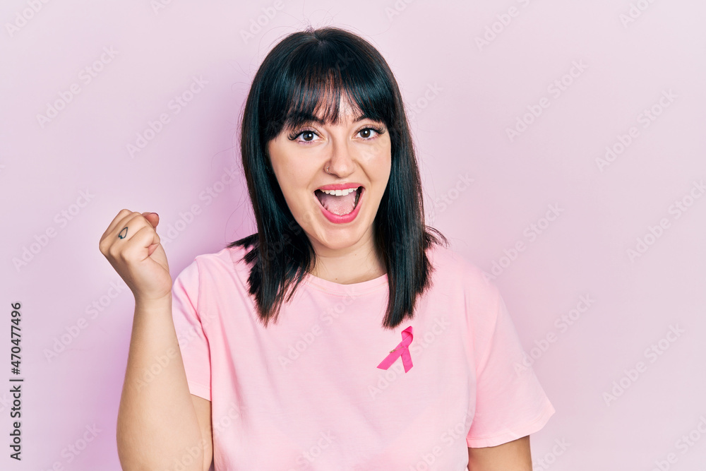 Young hispanic woman wearing pink cancer ribbon on shirt screaming proud, celebrating victory and success very excited with raised arms Young hispanic woman wearing pink cancer ribbon on shirt screaming proud, celebrating victory and success very excited with raised arms