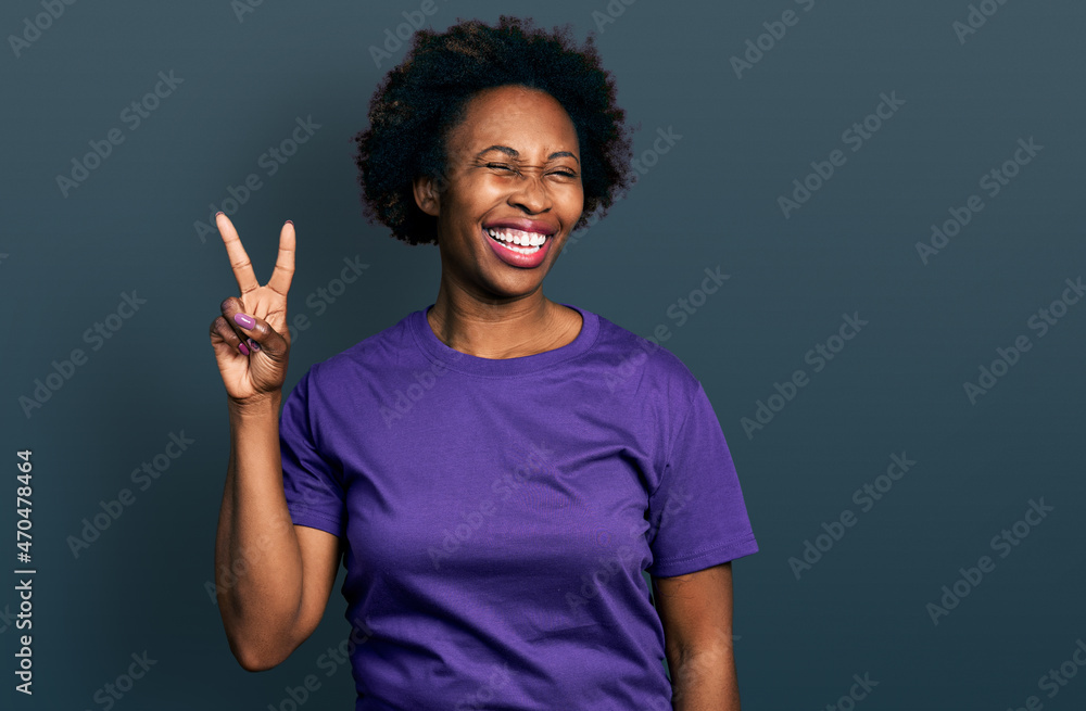 African american woman with afro hair wearing casual purple t shirt smiling with happy face winking at the camera doing victory sign with fingers. number two. African american woman with afro hair wearing casual purple t shirt smiling with happy face winking at the camera doing victory sign with fingers. number two.