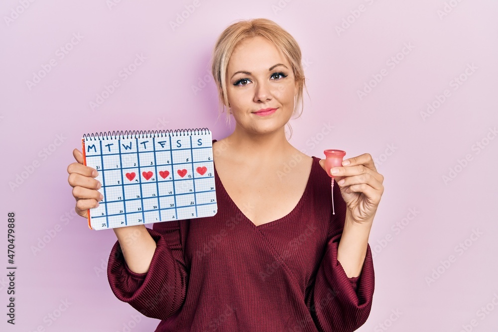 Young blonde woman holding heart calendar and menstrual cup relaxed with serious expression on face. simple and natural looking at the camera. Young blonde woman holding heart calendar and menstrual cup relaxed with serious expression on face. simple and natural looking at the camera.