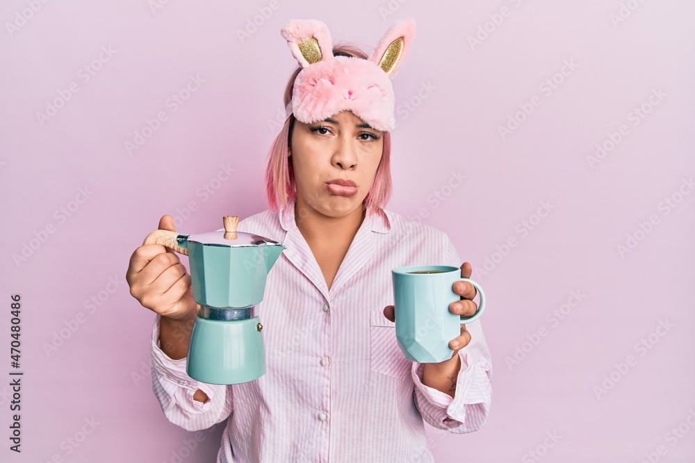Hispanic woman with pink hair wearing sleep mask and pajama drinking a coffee cup depressed and worry for distress, crying angry and afraid. sad expression. Hispanic woman with pink hair wearing sleep mask and pajama drinking a coffee cup depressed and worry for distress, crying angry and afraid. sad expression.