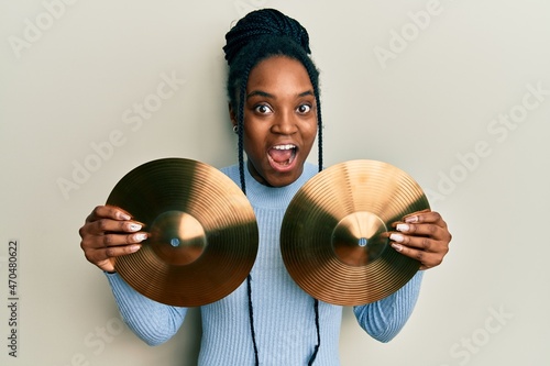 African american woman with braided hair holding golden cymbal plates celebrating crazy and amazed for success with open eyes screaming excited.